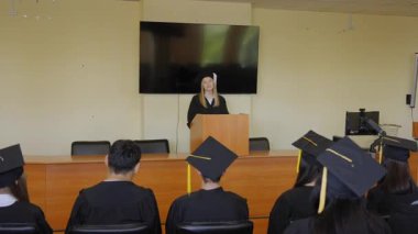 Genç bir kadın kürsüde duruyor ve sınıf arkadaşlarına mezuniyet konuşması yapıyor.