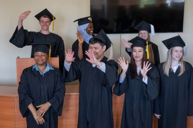 Mezuniyet elbisesi giymiş çok uluslu genç insanlar kürsüde duruyorlar.