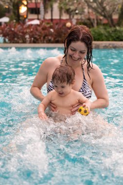 Beyaz bir kadın tatilde havuzda yüzerken oğlunu kucağına alıyor. Annesi küçük çocuğa yüzmeyi öğretiyor.