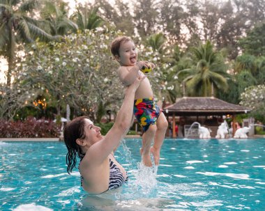 Beyaz bir kadın tatilde havuzda yüzerken oğlunu kucağına alıyor. Annesi küçük çocuğa yüzmeyi öğretiyor.