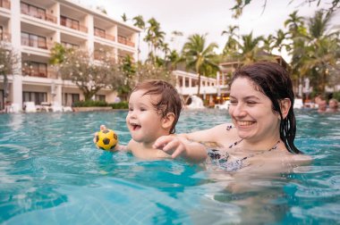 Beyaz bir kadın tatilde havuzda yüzerken oğlunu kucağına alıyor. Annesi küçük çocuğa yüzmeyi öğretiyor.