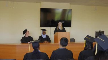 Genç bir kadın kürsüde duruyor ve sınıf arkadaşlarına mezuniyet konuşması yapıyor.