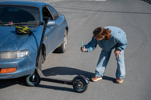 Woman examines and photographs the damage caused by a collision between an electric scooter and a car
