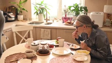 Orta yaşlı beyaz bir kadın oğluyla yemek yiyor.