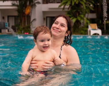 Beyaz bir kadın tatilde havuzda yüzerken oğlunu kucağına alıyor. Annesi küçük çocuğa yüzmeyi öğretiyor.