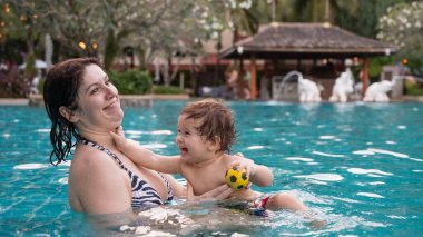 Beyaz bir kadın tatilde havuzda yüzerken oğlunu kucağına alıyor. Annesi küçük çocuğa yüzmeyi öğretiyor.