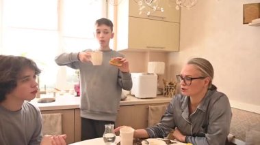 Orta yaşlı beyaz bir kadın iki oğluyla yemek yiyor.