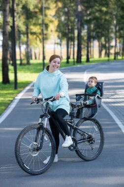 Beyaz bir kadın parkta bir yaşındaki oğluyla bisiklete biniyor. Dikey fotoğraf