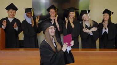 Sınıf arkadaşları onu alkışlarken, elinde diploma tutan beyaz bir kadın.