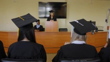 Genç bir kadın kürsüde duruyor ve sınıf arkadaşlarına mezuniyet konuşması yapıyor.