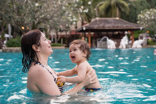 Beyaz bir kadın tatilde havuzda yüzerken oğlunu kucağına alıyor. Annesi küçük çocuğa yüzmeyi öğretiyor.