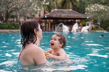 Beyaz bir kadın tatilde havuzda yüzerken oğlunu kucağına alıyor. Annesi küçük çocuğa yüzmeyi öğretiyor.