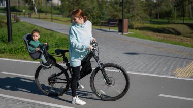 Beyaz kadın parkta bir yaşındaki oğluyla bisiklete biniyor.