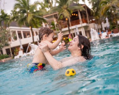Beyaz bir kadın tatilde havuzda yüzerken oğlunu kucağına alıyor. Annesi küçük çocuğa yüzmeyi öğretiyor.