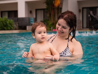 Beyaz bir kadın tatilde havuzda yüzerken oğlunu kucağına alıyor. Annesi küçük çocuğa yüzmeyi öğretiyor.