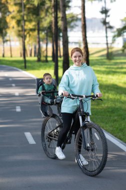 Beyaz bir kadın parkta bir yaşındaki oğluyla bisiklete biniyor. Dikey fotoğraf