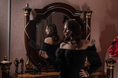 Siyah bir gece elbisesi içindeki güzel bir esmer eski tarz bir aynanın önünde poz veriyor.