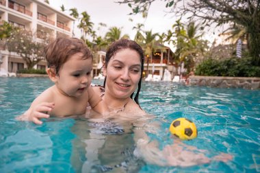 Beyaz bir kadın tatilde havuzda yüzerken oğlunu kucağına alıyor. Annesi küçük çocuğa yüzmeyi öğretiyor.