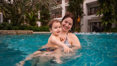 Beyaz bir kadın tatilde havuzda yüzerken oğlunu kucağına alıyor. Annesi küçük çocuğa yüzmeyi öğretiyor.