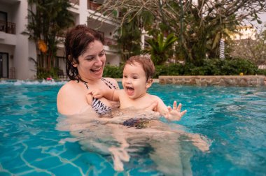 Beyaz bir kadın tatilde havuzda yüzerken oğlunu kucağına alıyor. Annesi küçük çocuğa yüzmeyi öğretiyor.