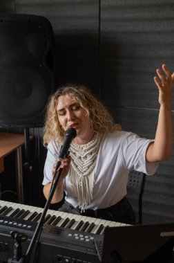 Beyaz kadın mikrofona şarkı söylüyor, sentezleyici çalıyor ve kendini dizüstü bilgisayarına kaydediyor.