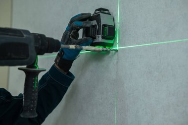 Bir adam lazer seviyesiyle mesafeyi ölçer ve çekiç matkabıyla duvarı deler.