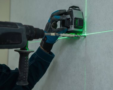 Bir adam lazer seviyesiyle mesafeyi ölçer ve çekiç matkabıyla duvarı deler.