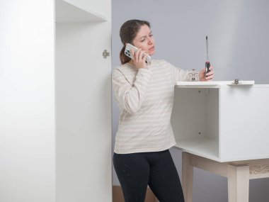 Beyaz bir kadın mobilya montajı yaparken telefonla konuşuyor.