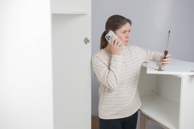 Beyaz bir kadın mobilya montajı yaparken telefonla konuşuyor.