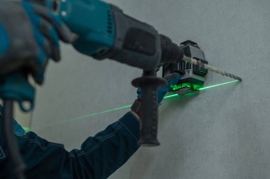 Bir adam lazer seviyesiyle mesafeyi ölçer ve çekiç matkabıyla duvarı deler.