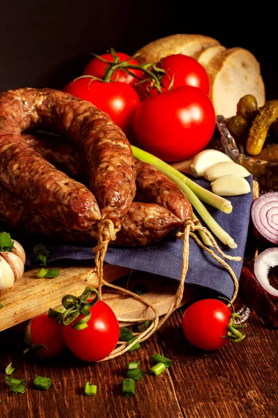 Appetizing smoked homemade sausage with herbs, fresh tomatoes, canned cucumbers, onions, bread and garlic — стоковое фото