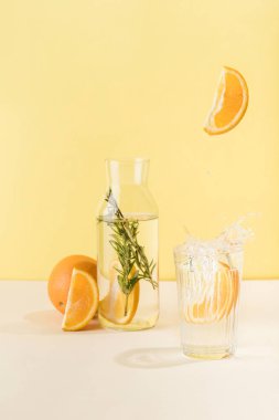 Bir bardak ve bir şişe temiz su. Yanında da portakal ve biberiye. Bir portakal bardağa bir damla suyla düşer. Metin için bir kopya alanı ile dikey yönelim.