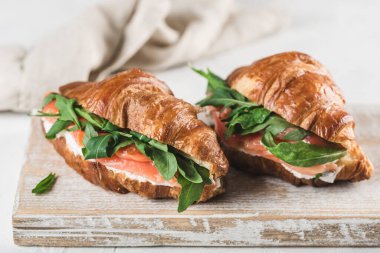 İki kruvasan sandviçi, somon balığı, ricotta peyniri ve arkaplanda peçete olan ahşap bir tahta üzerinde roka. Metin için bir kopya alanı ile yakın çekim. Yatay yönelim.