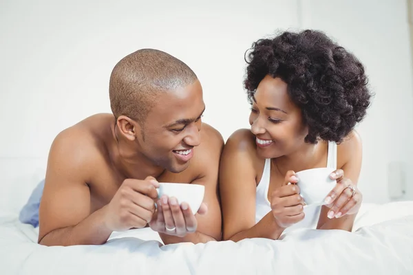 Happy couple lying on bed while holding cups — Stock fotografie