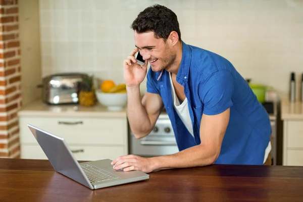 Hombre joven usando el ordenador portátil y hablando por teléfono —  Fotos de Stock