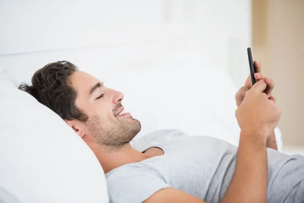 Мужчина улыбается, пользуясь мобильным телефоном на кровати — стоковое фото