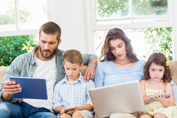 Rodina sedí na pohovce a pomocí notebooku, tabletu a telefonu — Stock fotografie
