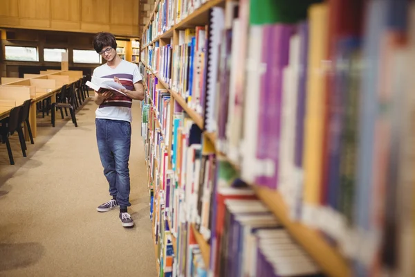 Молодой студент читает книгу, стоя возле книжной полки — стоковое фото