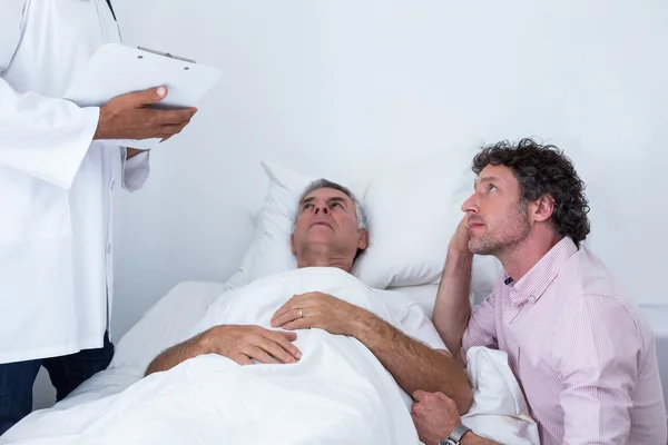 Мужчина, сидящий рядом с кроватью пациента и слушающий доктора — стоковое фото