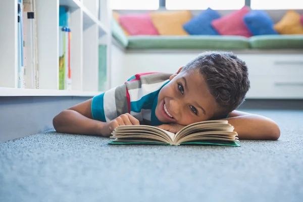 Skolpojke liggande på golvet och läsa en bok i biblioteket — Stockfoto