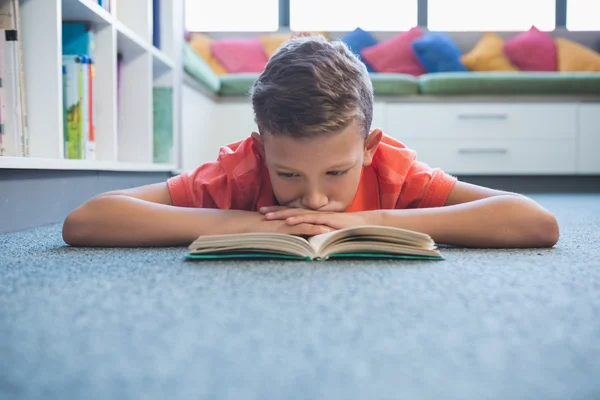 Colegial tumbado en el suelo y leyendo un libro en la biblioteca — Foto de Stock