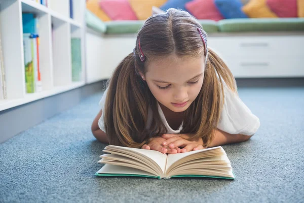 Skolflicka liggande på golvet och läsa en bok i biblioteket — Stockfoto