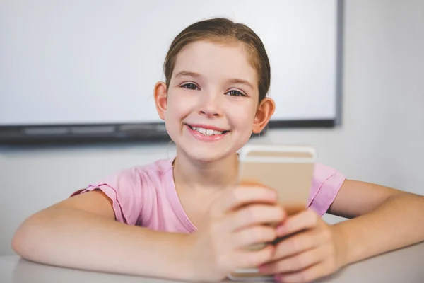 Smiling schoolgirl using mobile phone in classroom — Stock Photo, Image