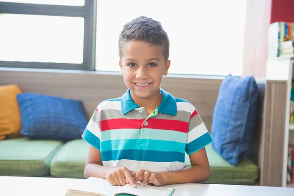Schoolboy sitting on table and reading book in library — Stock Photo, Image