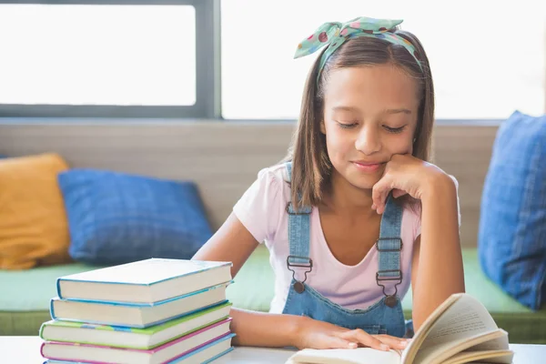 Colegiala sentada en la mesa y leyendo libro en la biblioteca —  Fotos de Stock