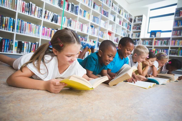 Miúdos da escola deitados no chão a ler livros na biblioteca — Fotografia de Stock