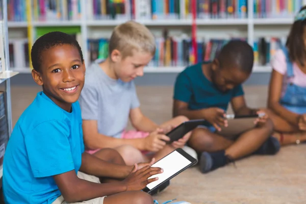 Niños de la escuela sentados en el suelo usando tableta digital en la biblioteca —  Fotos de Stock