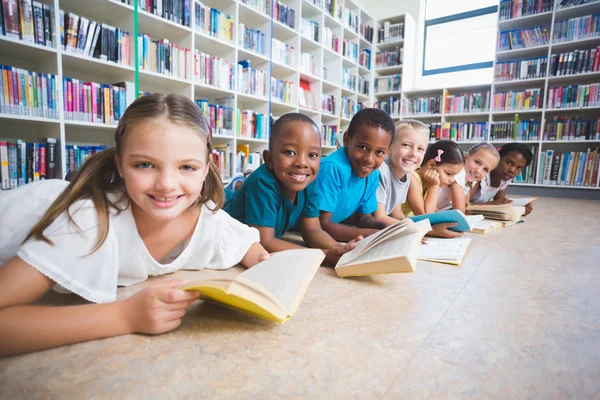 Niños sonrientes de la escuela tumbados en el piso leyendo un libro en la biblioteca —  Fotos de Stock