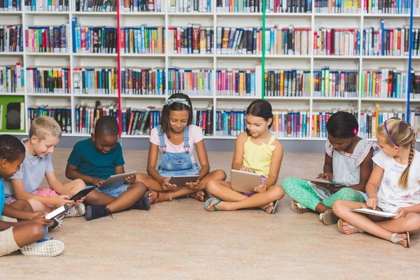 Niños de la escuela sentados en el suelo usando tableta digital en la biblioteca —  Fotos de Stock