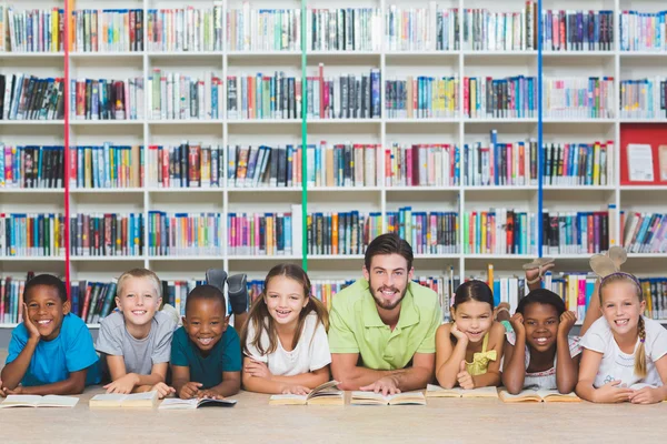 Profesor y niños tumbados en el suelo usando tableta digital en la biblioteca —  Fotos de Stock